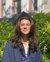 student stand in front of bushes, wearing a graduation gown