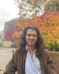 student with stripped shirt, jacket, and long hairs posing with a background of trees and buildings